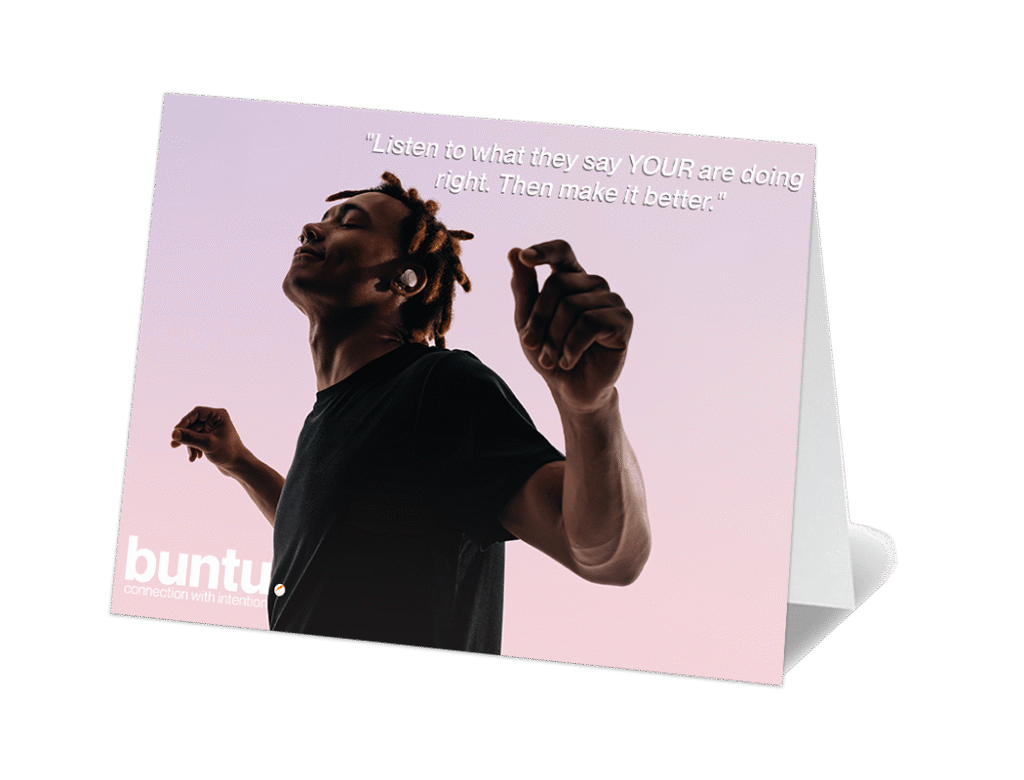 A greeting card stands upright, featuring an image of a person with dreadlocks looking upwards with a serene expression, arms slightly raised, set against a soft gradient sky of pink and purple. White text at the top reads: "Listen to what they say YOUR are doing right. Then make it better." The "buntu." logo is partially visible in the bottom left corner of the card.